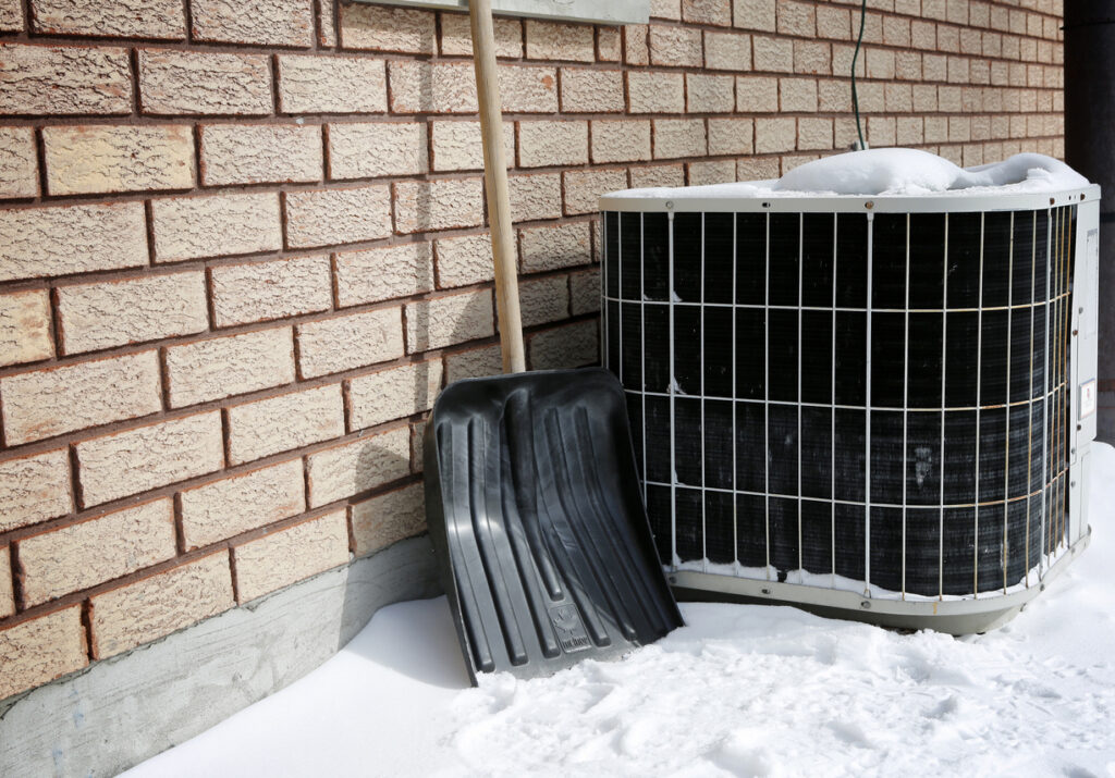Old outdoor HVAC system next to brick home; shovel leaning next to it with snow on the ground.