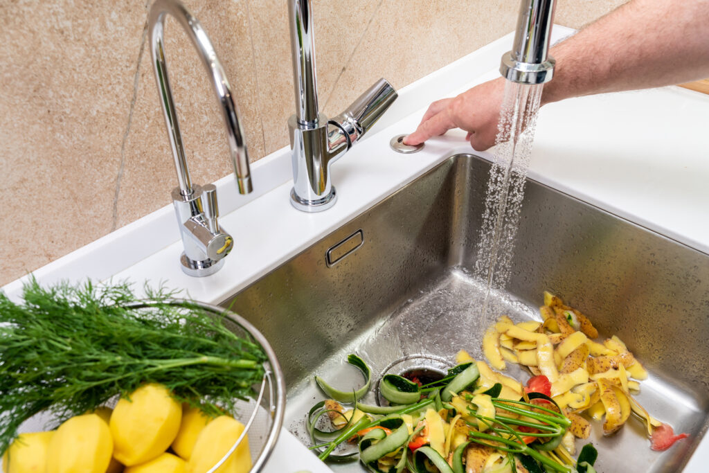 Close view of a sink full of vegetable scraps; a man presses a button to activate the garbage disposal.